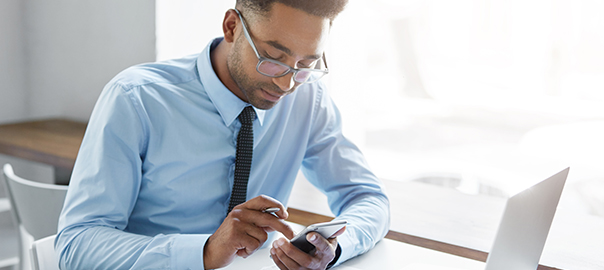 Picture of handsome African office worker having a few minutes break, texting sms on smart phone, sitting at his workplace with generic laptop computer, papers, notebook and mug of coffee on table