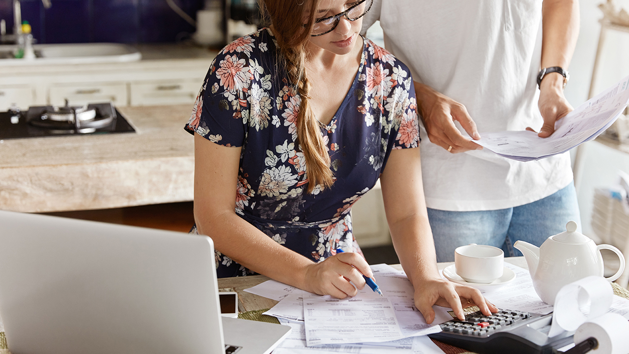 Cropped shot of pretty young woman calculates figures and writes down in papers, unrecognizable man stands near, helps with financial report. Domestic budget, family economy and expenses concept